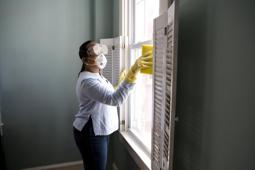 A woman cleans a window while wearing goggles and a medical mask