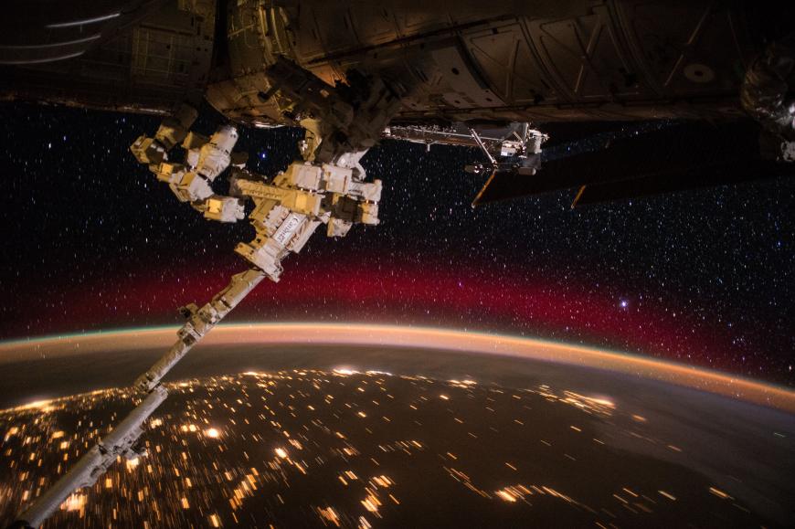 Photo from the ISS showing the exterior of the space station, the Canadarm, and Earth at night.
