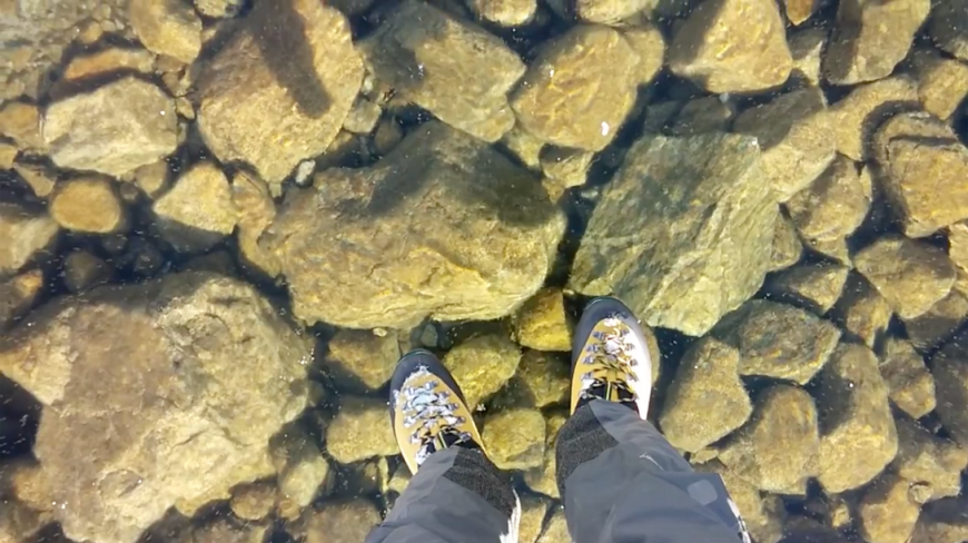 A photograph of a man&#039;s feet standing on a crystal clear frozen lake. The rocks are visible below.