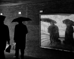 Black and white photo. Two men with umbrellas walk past a mirror