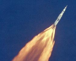 Apollo 11 Launch. A white rocketship against a dark blue sky with orange flames gushing from behind it.