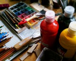 Art supplies spread out on a desk: paints in bottles, colored pencils, a paintbrush, water colors, a metal scaper