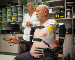 Man wears prosthetic arms with jointed fingers as researcher looks on.